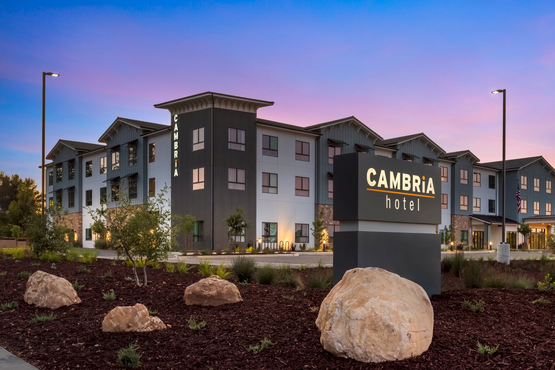 A Cambria Hotel building with a &ldquo;CAMBRIA hotel&rdquo; sign, vertical CAMBRIA on the facade, and lamp-lit exterior at dusk.