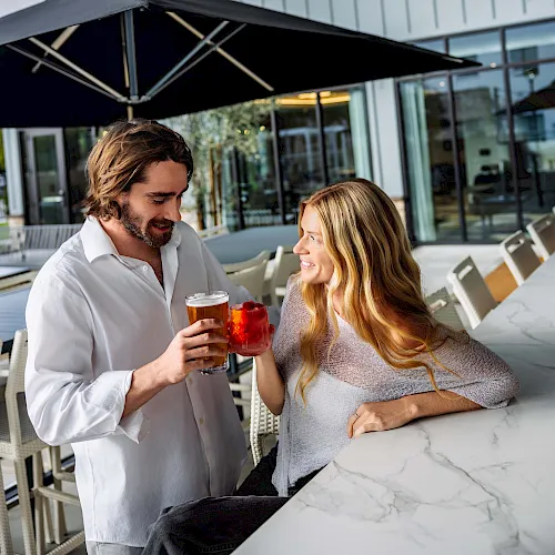 A man and woman sit on an outdoor patio under a dark umbrella, clinking drinks in the sunshine.