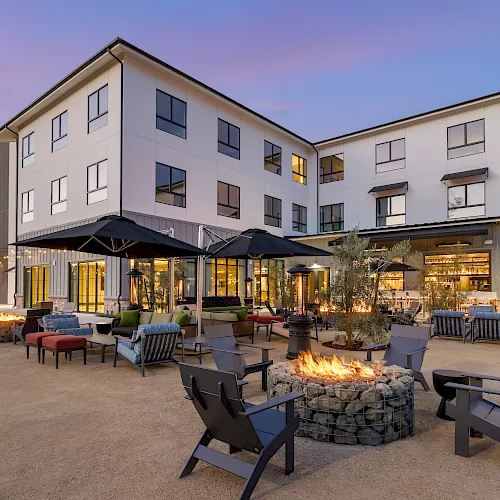 A modern apartment/hotel courtyard with outdoor seating, patio umbrellas, string lights, and a lit fire pit at dusk.