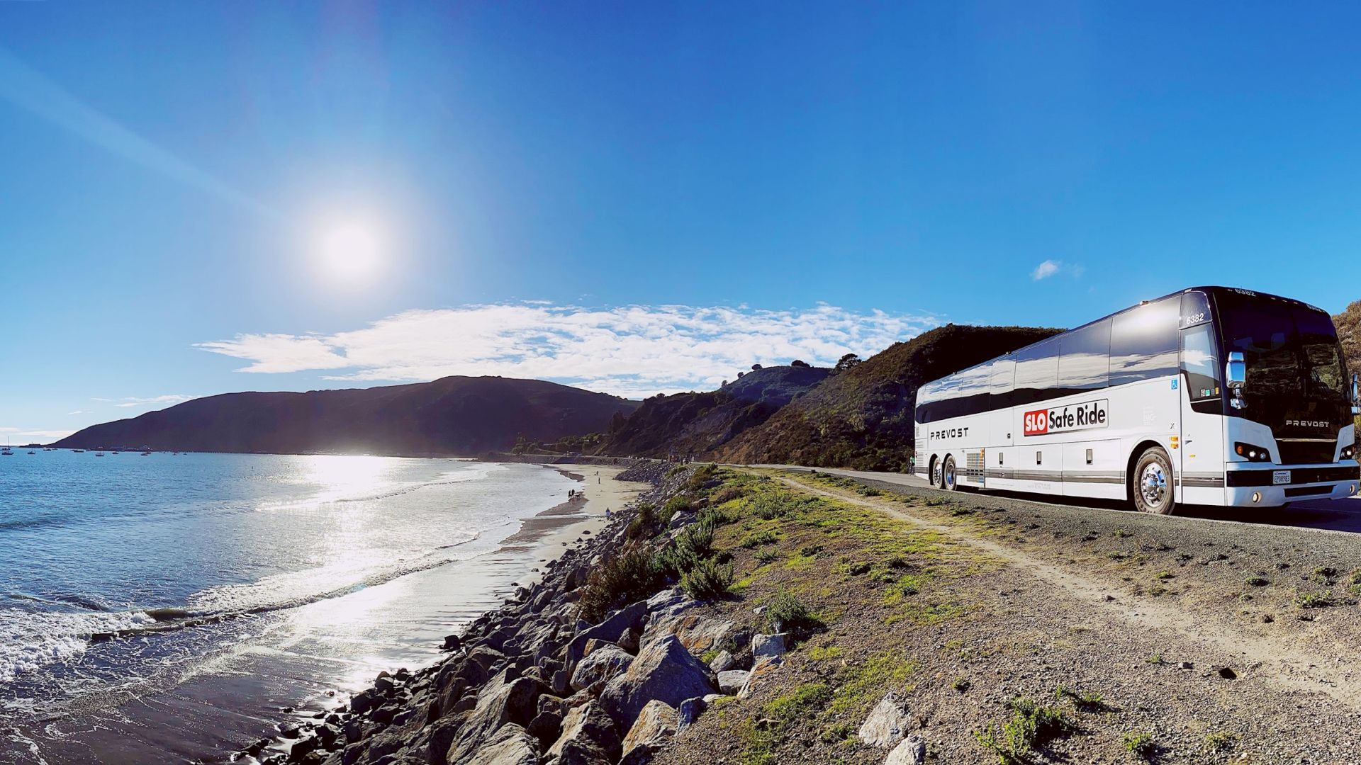 A white coach bus travels along a sunny coastal road beside a rocky beach and calm ocean under a bright blue sky.