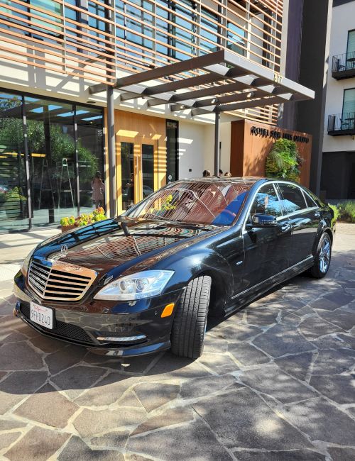A glossy black Mercedes sedan is parked under a modern awning outside a building, with colorful striped reflections on the hood.