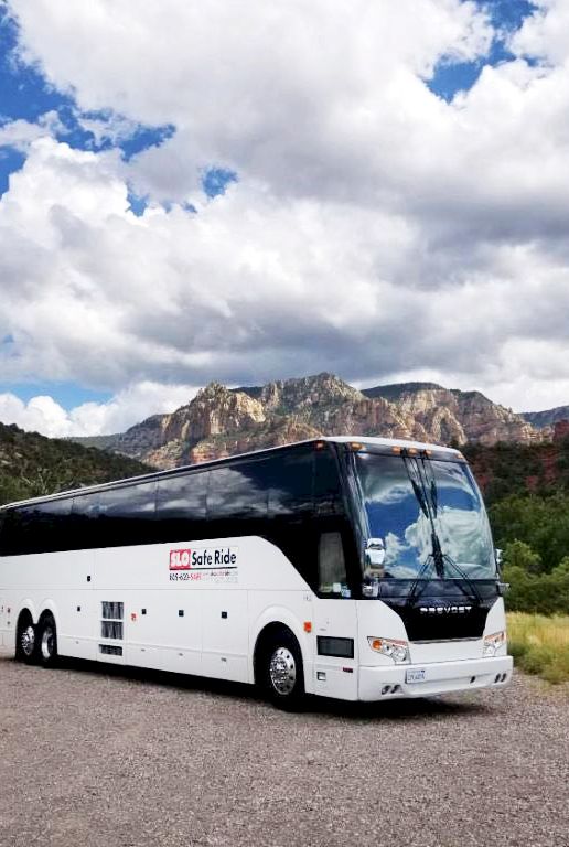 A white tour bus parked on a gravel road in a desert canyon, with red cliffs and cloudy skies behind it.
