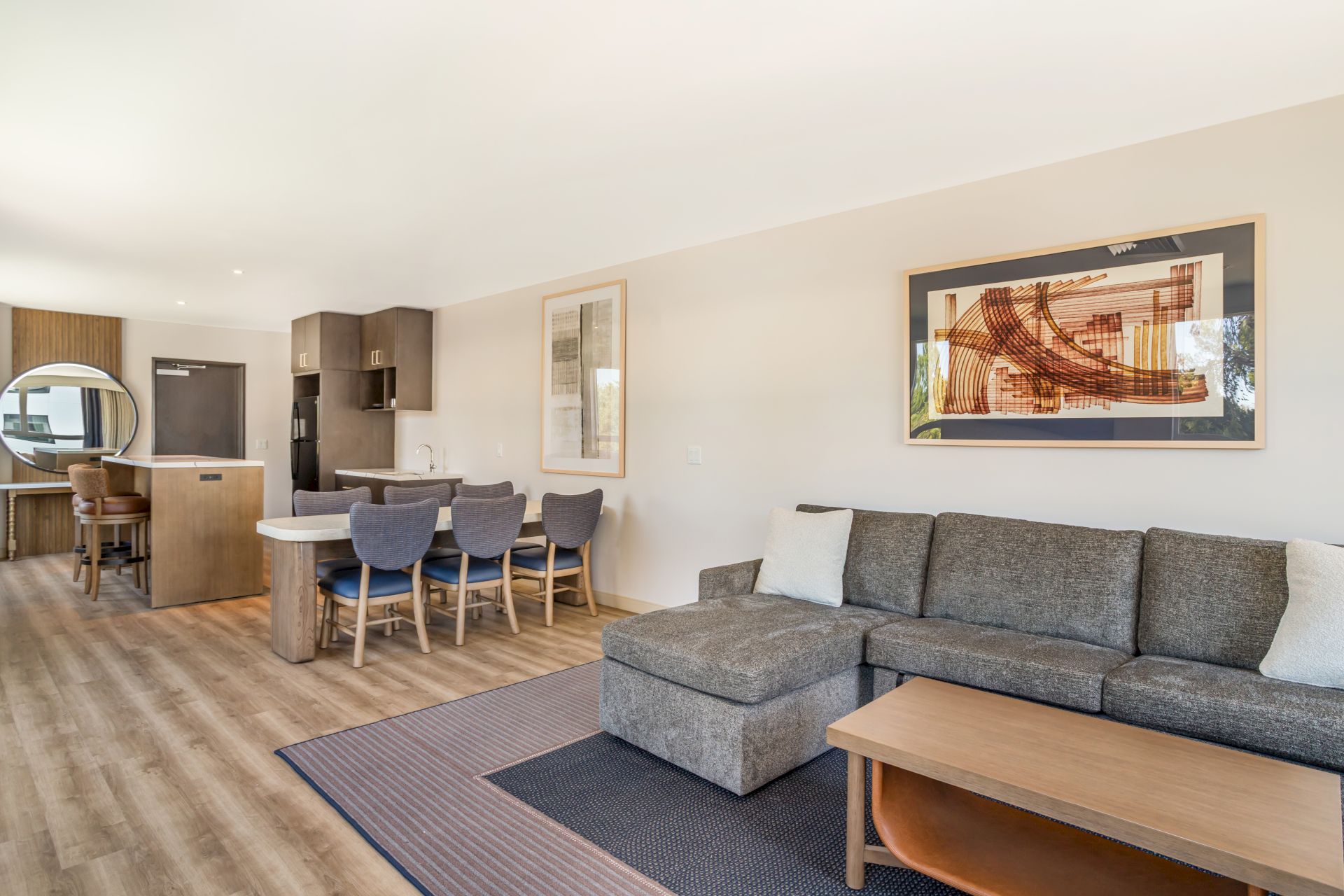 A modern living room with gray sofa, coffee table, dining set, and dark cabinetry kitchen with artwork.