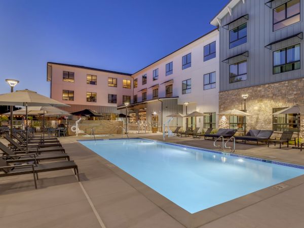 An outdoor hotel pool at dusk with sun loungers and umbrellas beside a modern multi-story building.