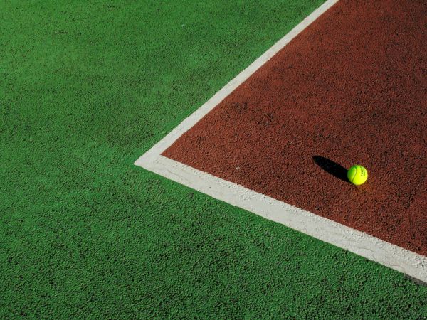 A green artificial field meets a red running lane, with a neon tennis ball resting near the white border.