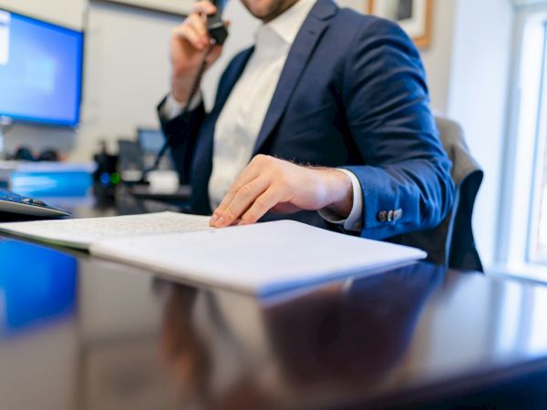 A suited office worker writes on documents while holding a phone, handling business tasks in a bright workplace.