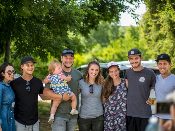 A group of friends and family pose outdoors in a green park beside a car, with one person holding a baby, smiling.