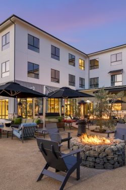 A modern hotel courtyard at dusk with patio furniture, multiple umbrellas, a fire pit, warm lights, and a bright multi-story building.