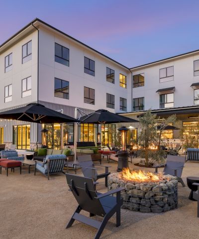 A modern hotel courtyard at dusk with patio furniture, multiple umbrellas, a fire pit, warm lights, and a bright multi-story building.