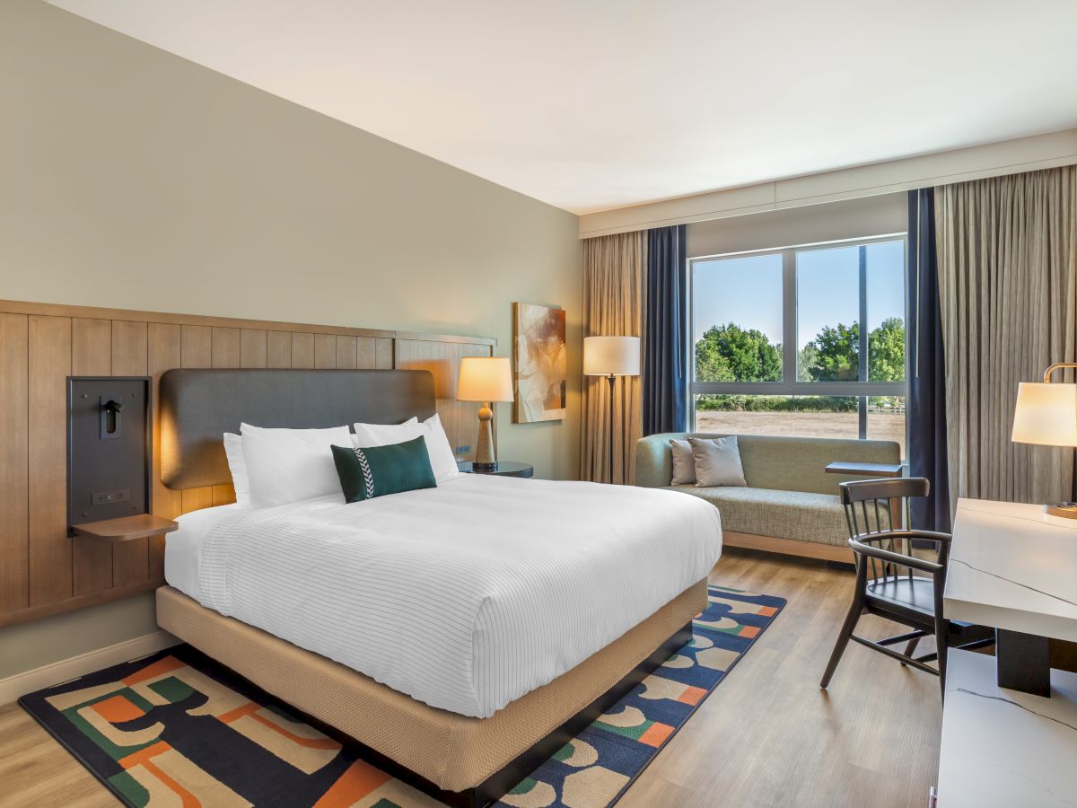 A modern hotel bedroom with a neatly made bed, bedside lamps, and a window showing greenery outside.