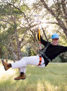 A zipline rider in helmet and harness whooshes through a forest, arms up, smiling, at a treetop adventure park.
