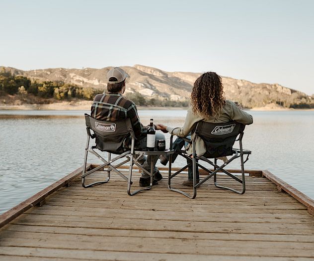 Two people sit in Coleman folding chairs on a wooden dock, enjoying a lakeside view of mountains in the evening light.