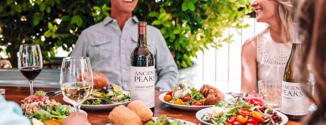 An outdoor dinner with two smiling friends, plates of salad and bread, and &ldquo;Ancient Peaks&rdquo; wine bottles on a wooden table.