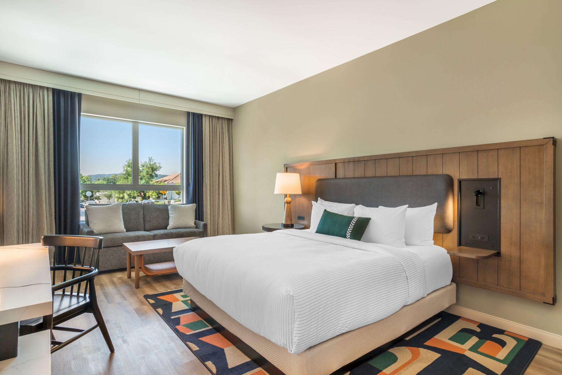 A modern hotel room with a neatly made bed, bedside lamp, curtains, and a windowed seating area.