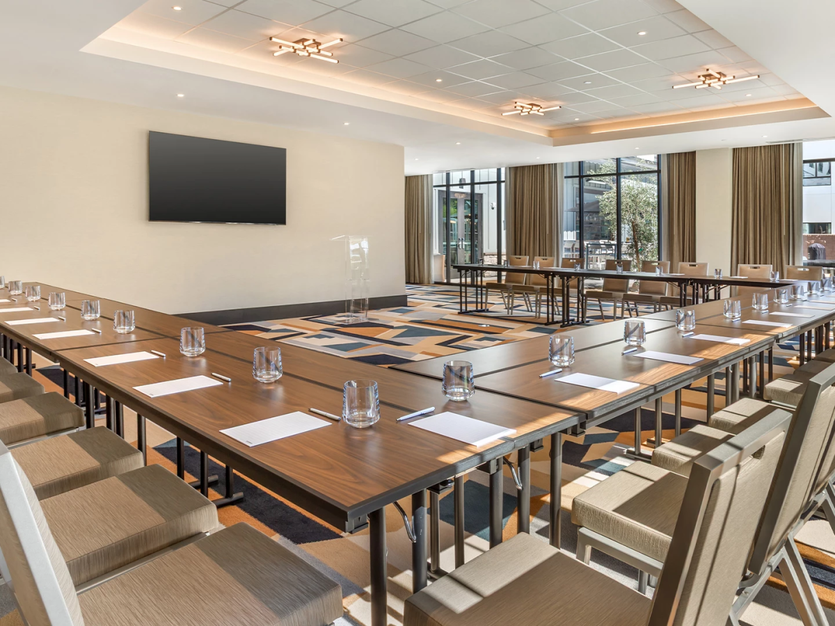 A modern conference room with long wooden tables set with glassware, chairs, and a large TV, lit by ceiling chandeliers.