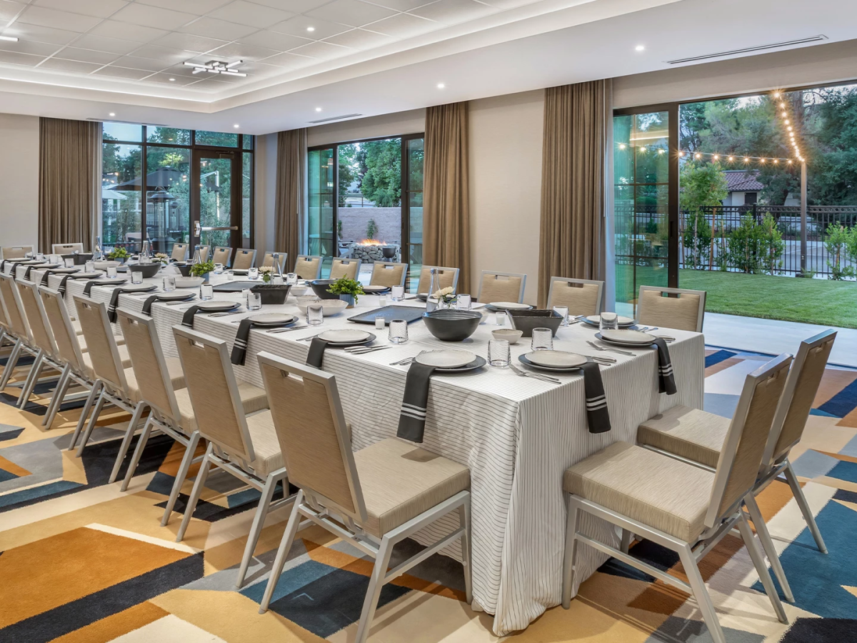 An elegant indoor dining room with a long set table, modern chairs, large glass doors, and warm string lights outside.