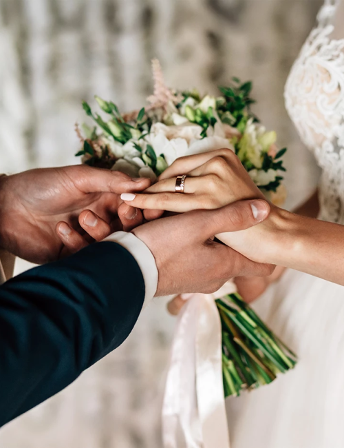 A couple exchanging rings with joined hands, holding a wedding bouquet; bridal lace and groom suit in a soft romantic scene.