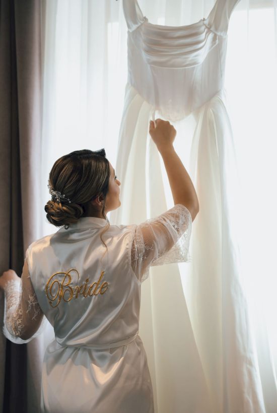 A bride in satin robe lifts her wedding dress/veil in bright morning light by a window, getting ready for her big day.