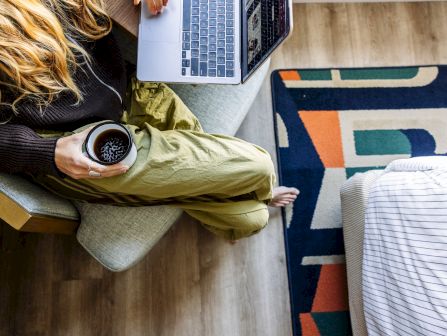 A woman lounges on a couch with a laptop above her, holding a cup of coffee, while patterned blankets cover her legs.
