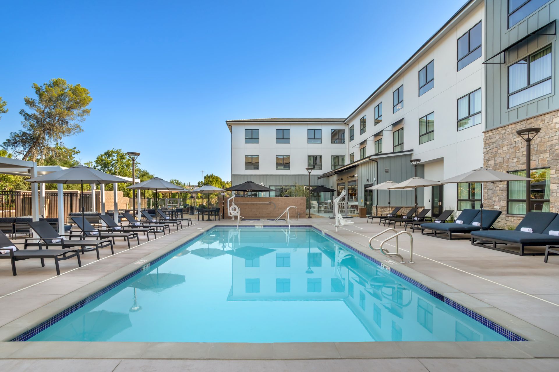 Outdoor hotel courtyard with a blue swimming pool, lounge chairs, umbrellas, and a modern multi-story building under a clear sky.