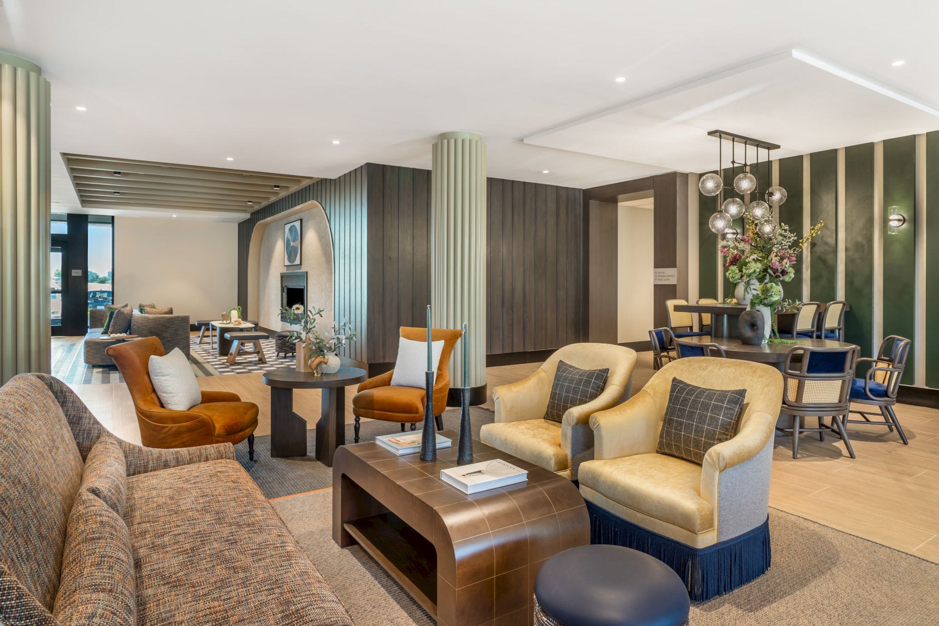 A modern lounge and dining area with plush sofas, a coffee table, wood-paneled walls, and a sparkling globe-chandelier above flowers.
