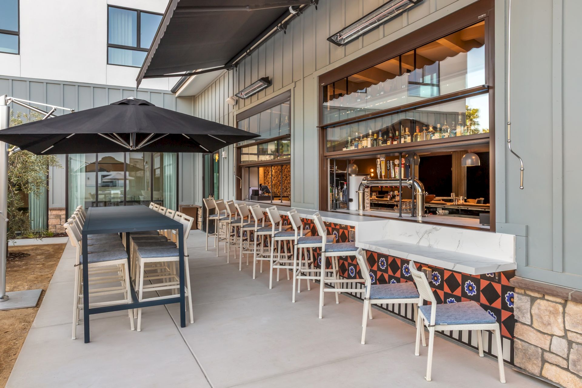 Outdoor patio with tables, chairs, black umbrellas, and a service bar stocked with bottles and drinks in warm light.