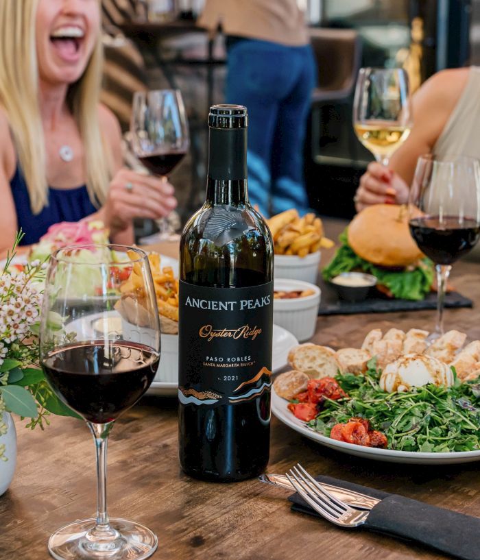 A wine bottle and glasses sit on a table during a lively dinner with friends, plates of food, and cheerful laughter.