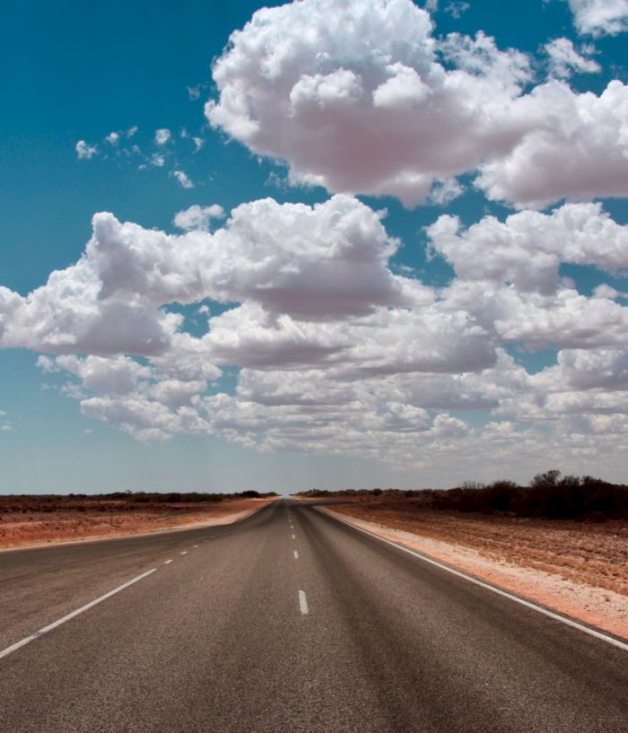 A long desert road stretches toward towering clouds in a bright blue sky.