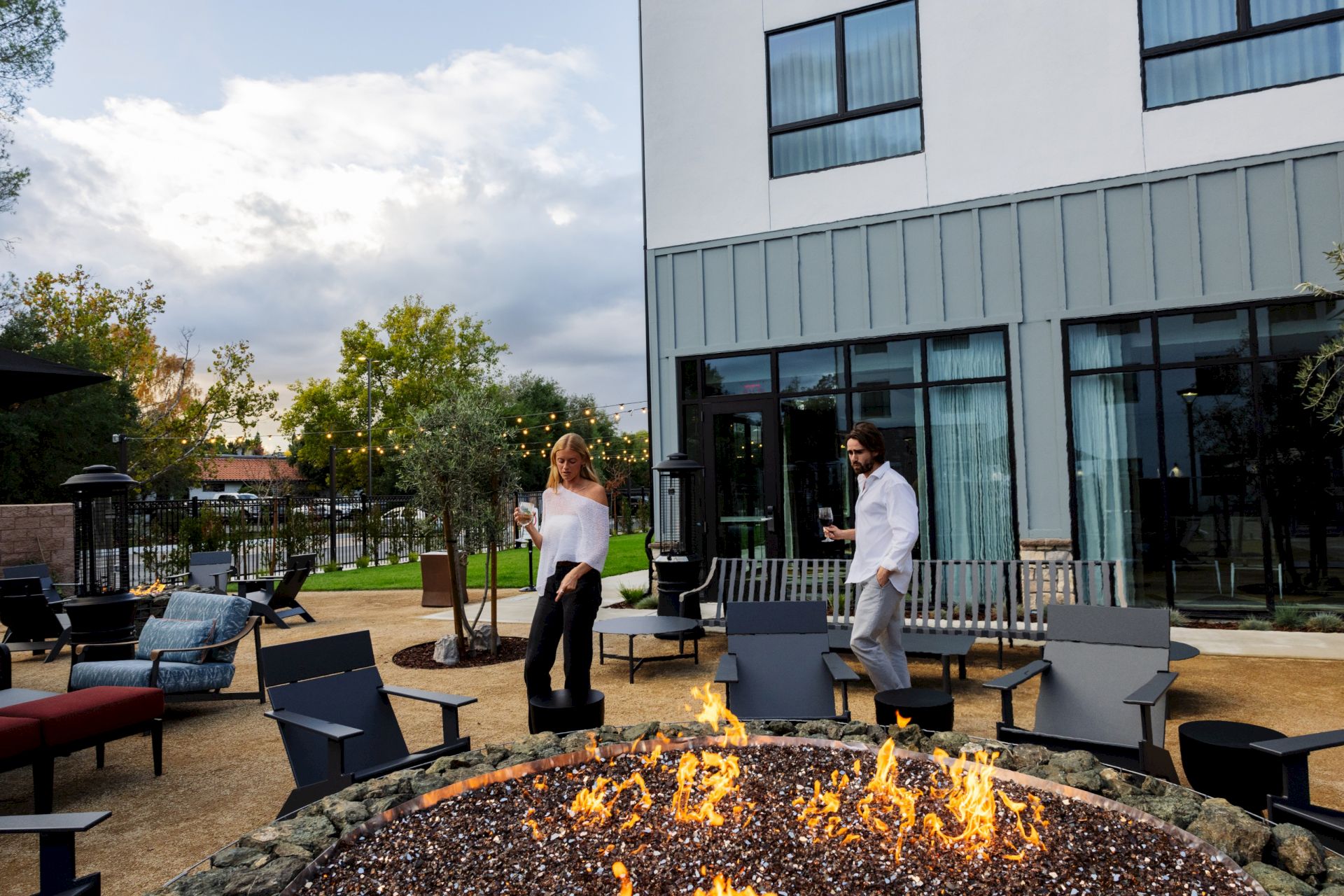 An outdoor patio by a blazing fire pit, where two people hold drinks near a modern building and twinkling lights.