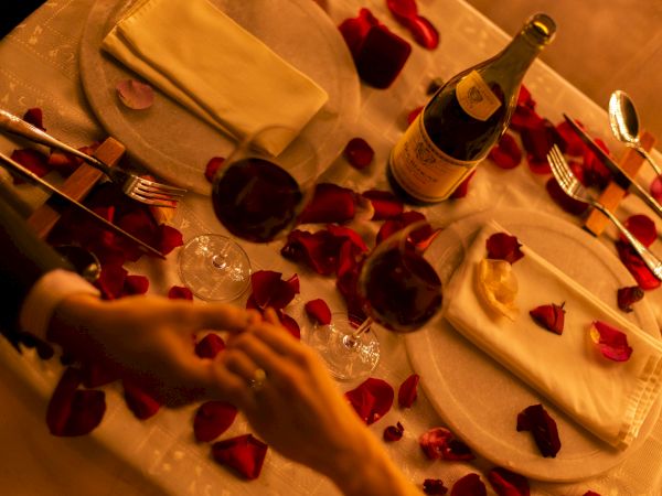 A romantic table setting with wine bottles and glasses, rose petals, and folded napkins with cutlery on white linens.