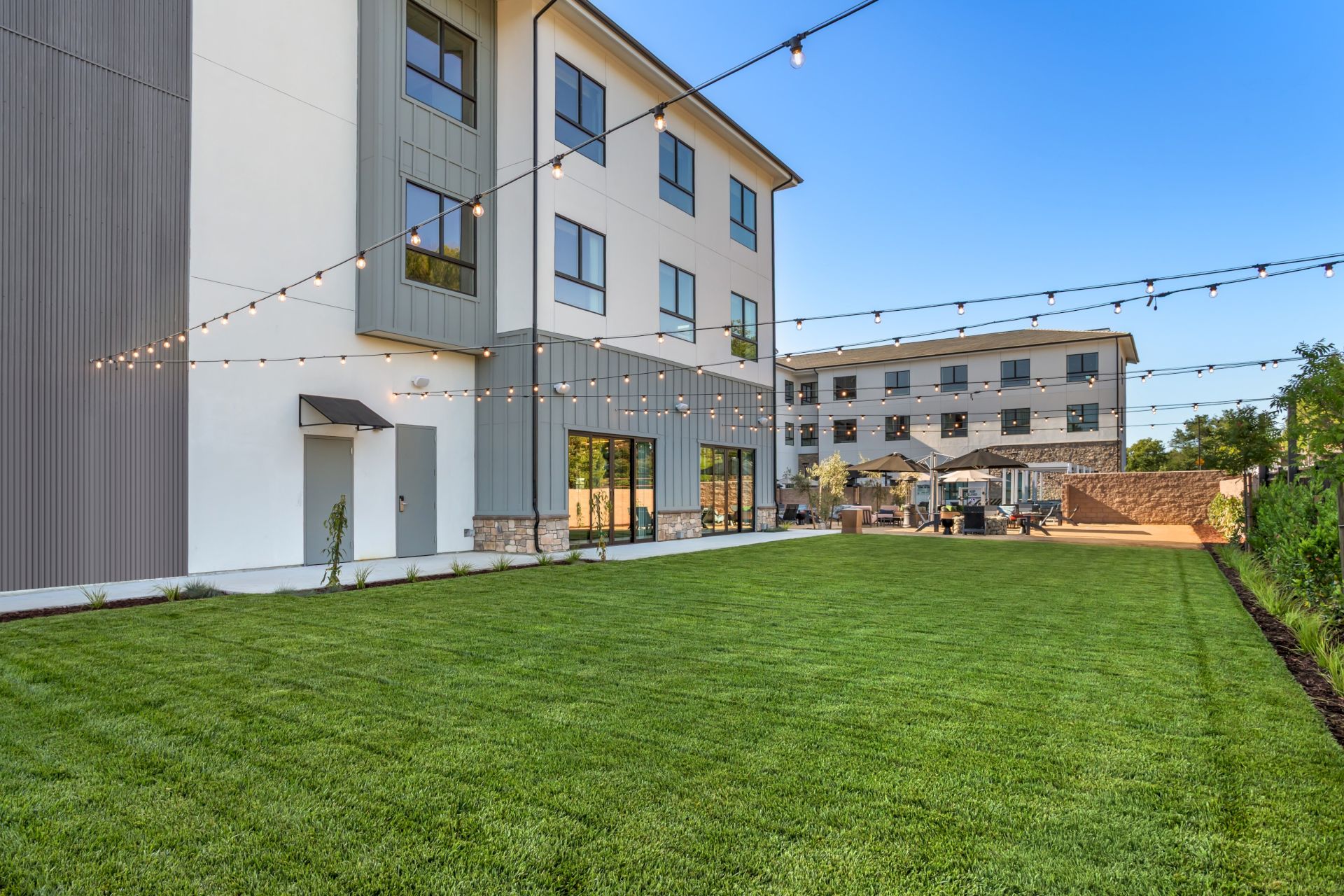 A modern building with a green lawn, outdoor patio seating, and warm string lights strung overhead for evening gatherings.