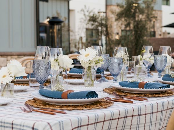 An outdoor dining table set with ornate glassware, blue napkins, and white daisies, styled for a celebration or wedding reception.