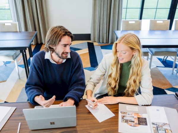 A man and woman sit at a table discussing plans with laptops and papers in a bright office.