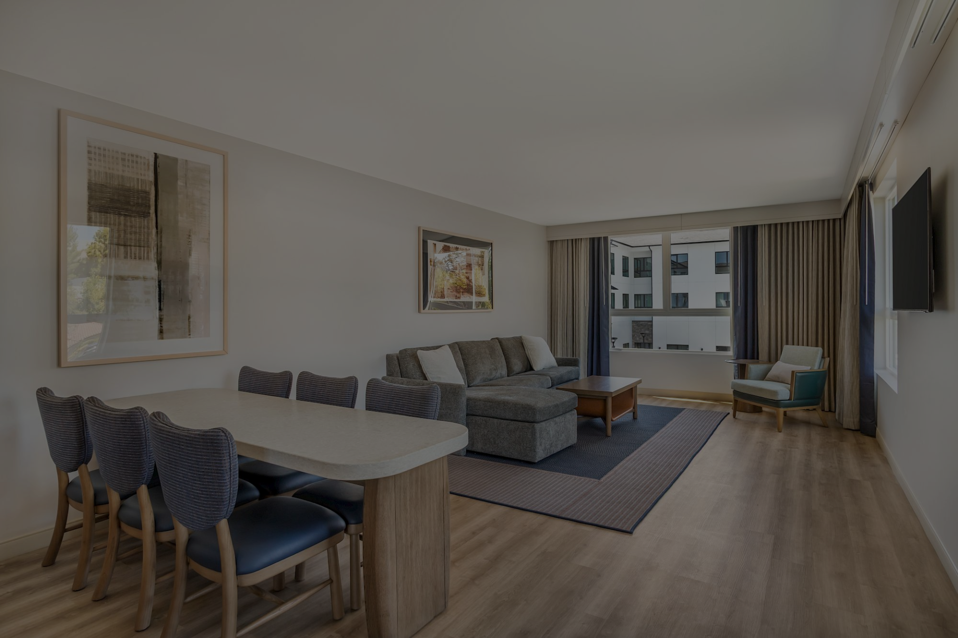A bright modern living room with a grey sofa, dining table, TV, curtains, and large windows overlooking nearby buildings.