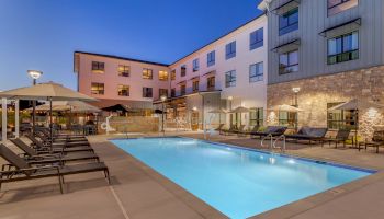 A hotel courtyard with a lit swimming pool, loungers, umbrellas, and modern multi-story building at dusk.